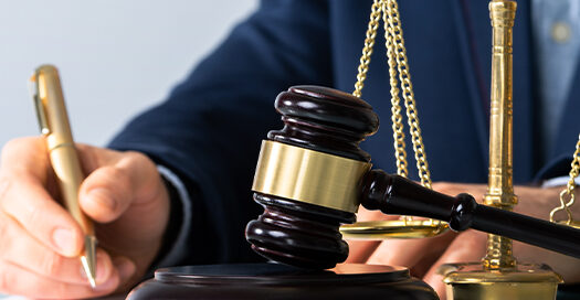 A man in a suit writes on a document alongside a gavel and metal scales of justice.