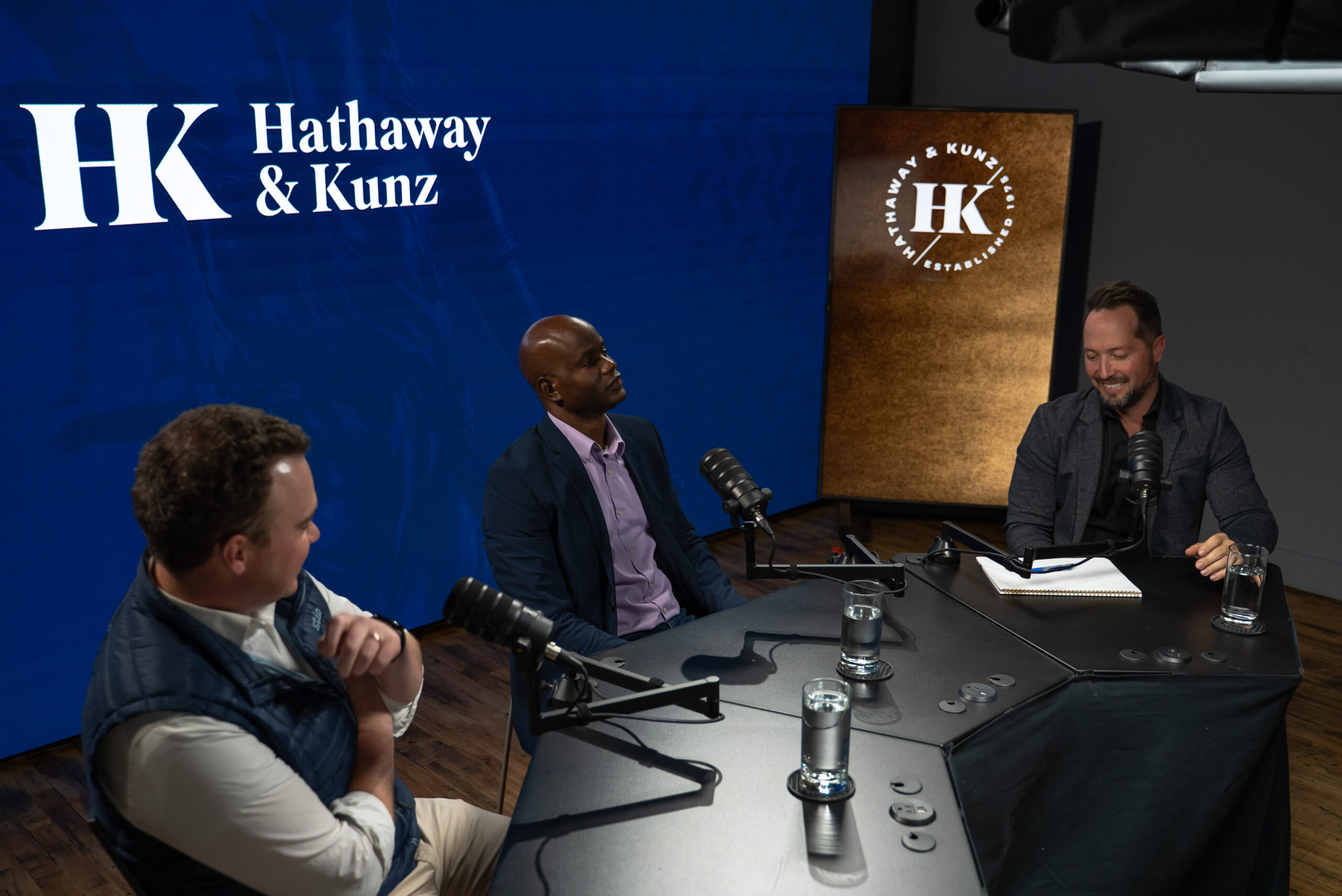Hathaway_BTS_Podcast4 Three males sit around a table, recording a podcast.
