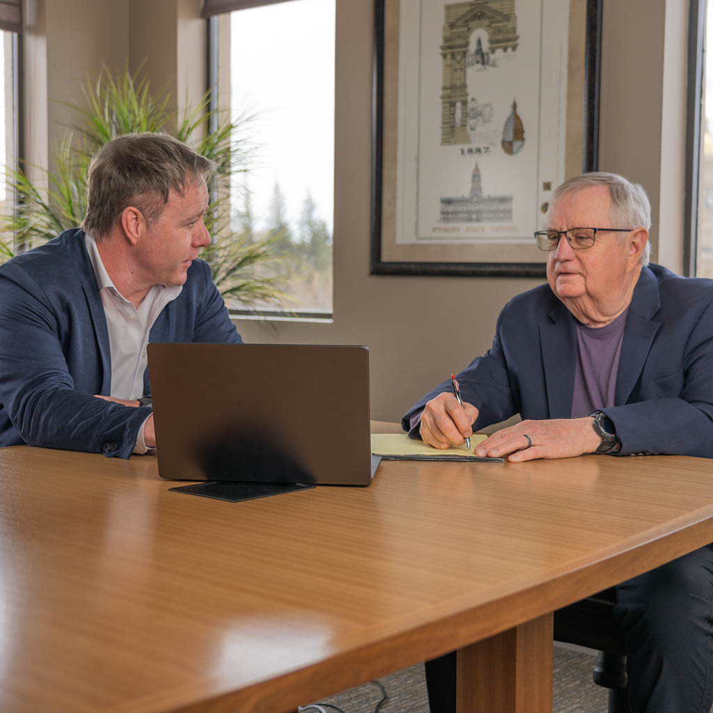 Two men at at a table, reviewing information on a laptop.